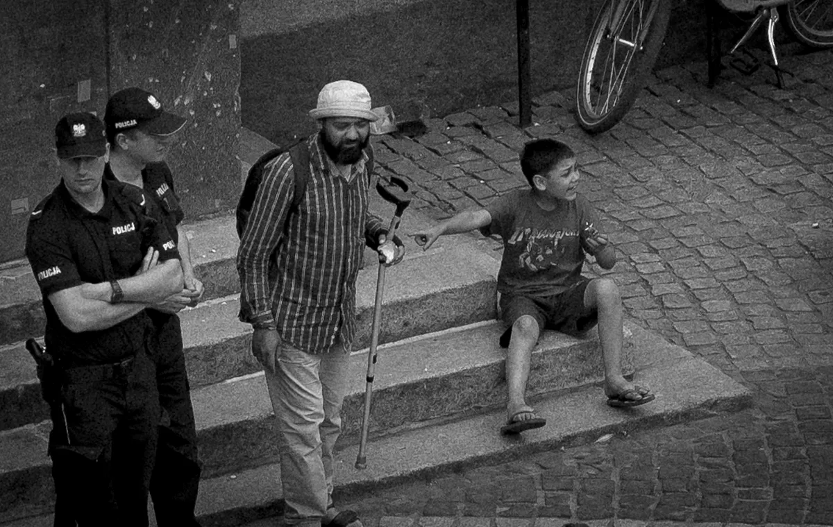 Two police officers beside a bearded man with a crutch while a seated boy points away